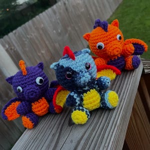 May include: Three handmade crochet dragon toys in purple, orange, and blue. Each dragon has large googly eyes, wings, and a small horn. The dragons are sitting on a wooden surface.