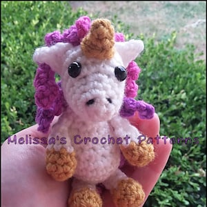 Puede incluir: Un unicornio de crochet blanco con un cuerno dorado y una crin morada. El unicornio está sosteniendo sus pezuñas en el aire.