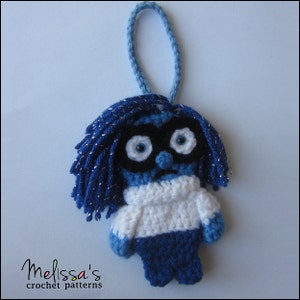 May include: A blue and white crocheted ornament of a character with blue hair, a white sweater, and a sad expression. The ornament has a blue loop for hanging.