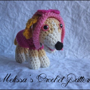 Op de afbeelding: Een gehaakt hondenspeelgoed van roze en geel garen, met een roze strik. Het speelgoed heeft bruine ogen en een zwarte neus.