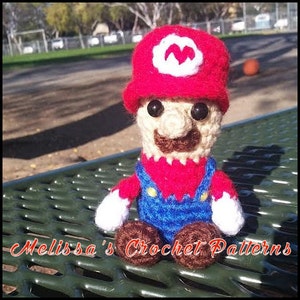May include: A crocheted Mario character wearing a red hat with a white 'M' on it, a red shirt, blue overalls, and brown shoes. The character is sitting on a green metal surface.