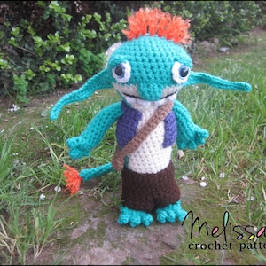 Può includere: Una bambola all'uncinetto fatta a mano con un design stravagante. La bambola presenta una testa e braccia turchesi, capelli arancioni e una borsa marrone. Indossa un gilet viola e pantaloncini marroni. La bambola è in piedi su una superficie di pietra con erba sullo sfondo. Il testo "Melissa's crochet patterns" è visibile.