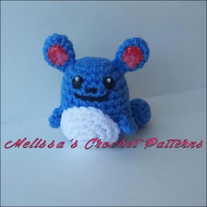 May include: A blue and white crocheted plush toy with red ears. The toy has a smiling face with black eyes and a black nose. The toy is sitting on a white surface. Melissa's Crochet Patterns.