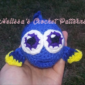 May include: A crocheted blue fish with yellow and white accents. The fish has large purple and white eyes. The fish is being held in a hand. "Melissa's Crochet Patterns" is written in the background.