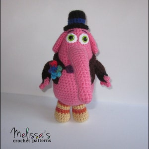 May include: A pink crocheted elephant with a brown jacket, a black top hat with a blue band, and green eyes. The elephant is holding a colorful crocheted flower.