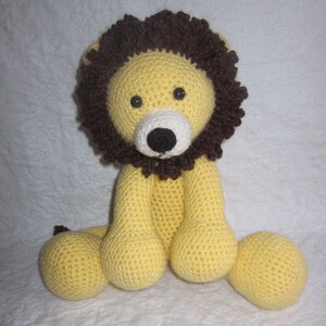 May include: A crocheted stuffed lion toy with a yellow body, brown mane, and black eyes. The lion is sitting with its legs crossed and its tail curled behind it.