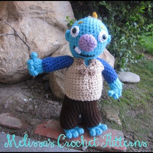 Puede incluir: Un juguete monstruo de crochet azul y marrón con una nariz morada y un palo en la mano. El juguete está de pie sobre una superficie de ladrillo.