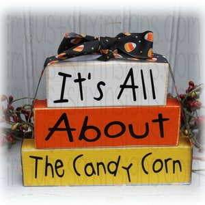 May include: Stacked wooden blocks with the words "It's All About The Candy Corn" painted on them. The blocks are decorated with a black and white candy corn ribbon.