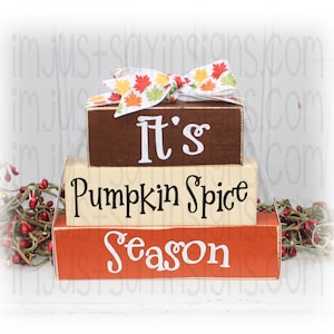 May include: A three-tiered wooden sign with the words "It's Pumpkin Spice Season" in black lettering. The top tier is brown, the middle tier is cream, and the bottom tier is orange. The sign is decorated with a fall-themed ribbon.