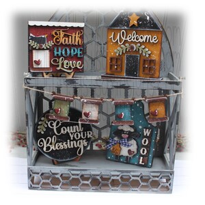 May include: A decorative metal shelf displays several wooden signs. One sign reads "Faith Hope Love" in script, another says "Welcome" on a house-shaped sign. A garland of spools hangs below, and a sign with a sheep reads "Wool".