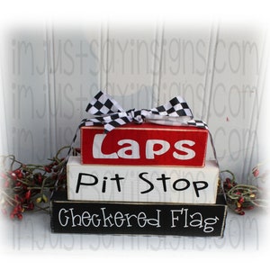 May include: Three wooden blocks stacked on top of each other. The top block is red and says "Laps" in white letters. The middle block is white and says "Pit Stop" in black letters. The bottom block is black and says "Checkered Flag" in white letters. A black and white checkered ribbon is tied around the top block.