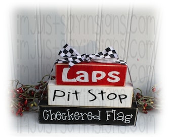 May include: Three wooden blocks stacked on top of each other. The top block is red and says "Laps" in white letters. The middle block is white and says "Pit Stop" in black letters. The bottom block is black and says "Checkered Flag" in white letters. A black and white checkered ribbon is tied around the top block.