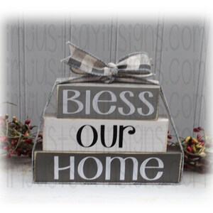 May include: A three-tiered wooden block sign with the words "Bless Our Home" in white and black lettering. The blocks are painted gray and white, and a black and white checkered ribbon is tied around the top block.