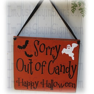 May include: A rectangular orange wooden sign with a black ribbon hanger. The sign features black text that reads Sorry Out of Candy and Happy Halloween, accompanied by black silhouettes of two bats and a white ghost graphic on the right side.