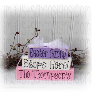 May include: Three wooden blocks stacked on top of each other. The top block is lavender and says "Easter Bunny". The middle block is white and says "Stops Here!" The bottom block is pink and says "The Thompson's".