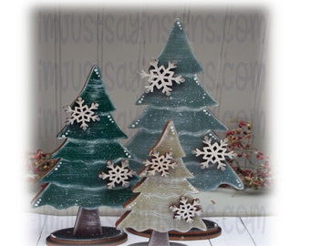 May include: Set of three wooden Christmas trees in varying sizes. The trees are painted in shades of green and gray, with white accents and wooden snowflake ornaments. Each tree stands on a round, dark brown base, creating a rustic holiday decoration.