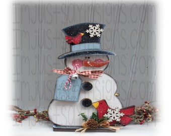 May include: A wooden snowman figurine with a blue and white top hat, a red cardinal, and a tag that says "Let it Snow". The snowman is holding a red cardinal in its right hand. The snowman is standing on a wooden base with a pine cone and straw.