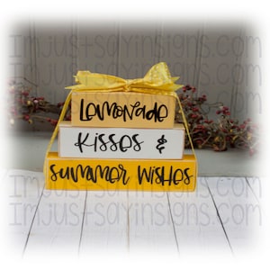May include: Stacked wooden blocks with yellow and white paint. The top block says "Lemonade", the middle block says "Kisses &" and the bottom block says "Summer Wishes".