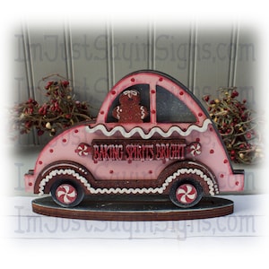May include: A pink gingerbread car with a gingerbread person in the driver's seat. The car has candy cane wheels and a sign that reads "Baking Spirits Bright".