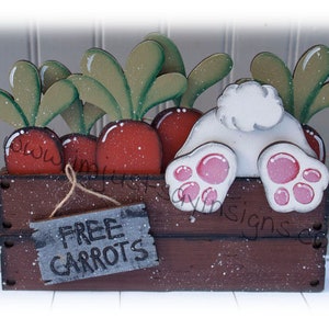 May include: A wooden bunny rabbit figurine with a white tail and pink feet sits in a brown wooden crate with a sign that reads "FREE CARROTS". The crate is filled with painted carrots with green tops.