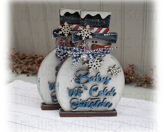 Pode incluir: Duas figuras de boneco de neve de madeira com chapéus pretos, cachecóis listrados vermelho e branco e detalhes de flocos de neve azuis e brancos. O boneco de neve à direita tem um fundo branco com o texto "Baby It's Cold Outside" em letras azuis e brancas.