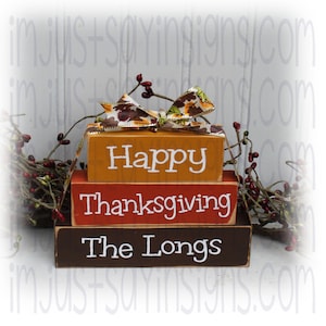 May include: A three-tiered wooden block sign with the words "Happy Thanksgiving The Longs" painted in white. The top block is orange, the middle block is red, and the bottom block is brown. A fall-themed ribbon with leaves is tied around the top block.