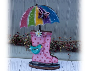May include: A wooden rain boot with pink polka dots and a blue bird perched on the side. A colorful umbrella with a butterfly sits on top of the boot. The boot is on a wooden base.
