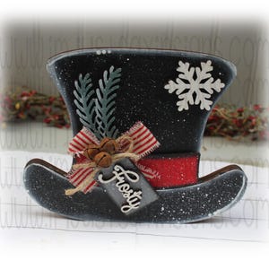 May include: A black and white snowman hat with a red band, a white snowflake, and a green sprig of holly. The hat has a tag that says "Frosty".