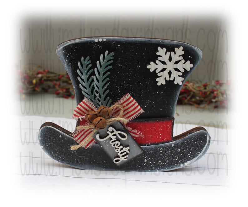 May include: A black and white snowman hat with a red band, a white snowflake, and a green sprig of holly. The hat has a tag that says "Frosty".