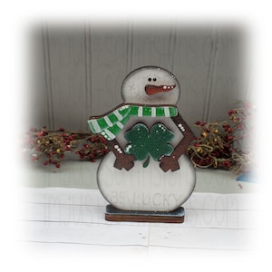May include: A wooden snowman decoration with a green and white scarf and a shamrock. The snowman has a carrot nose and the words "BE LUCKY" are printed on the base. The snowman is approximately 6 inches tall.