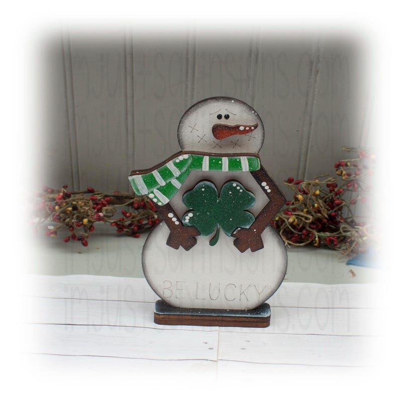 May include: A wooden snowman decoration with a green and white scarf and a shamrock. The snowman has a carrot nose and the words "BE LUCKY" are printed on the base. The snowman is approximately 6 inches tall.