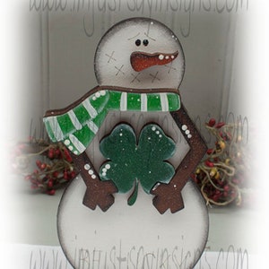 May include: Wooden snowman decoration with a green and white striped scarf and a green shamrock. The snowman has a carrot nose and a painted face. The base is a dark blue color. The website address www.imjustsayinsigns.com is printed on the image.