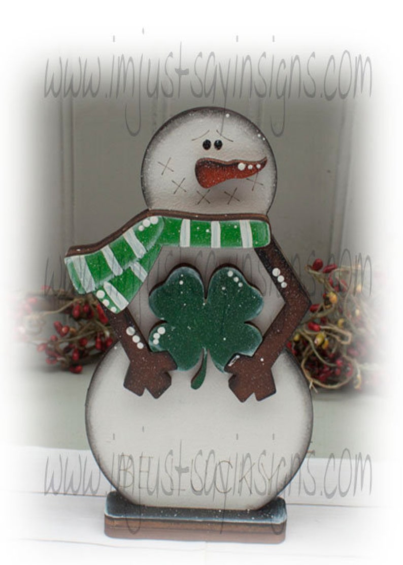 May include: Wooden snowman decoration with a green and white striped scarf and a green shamrock. The snowman has a carrot nose and a painted face. The base is a dark blue color. The website address www.imjustsayinsigns.com is printed on the image.