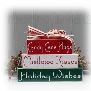 May include: A stack of three wooden blocks with holiday-themed messages. The top block is red with "Candy Cane Hugs," the middle is white with "Mistletoe Kisses," and the bottom is green with "Holiday Wishes." A red and white striped bow adorns the top.