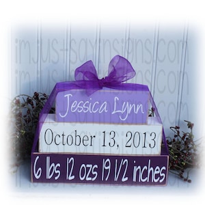 May include: Purple, white, and lavender wooden blocks stacked on top of each other. The top block says "Jessica Lynn", the middle block says "October 13, 2013", and the bottom block says "6 lbs 12 ozs 19 1/2 inches".