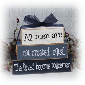 May include: Stacked wooden blocks with a navy blue ribbon. The top block is white with the text "All men are" in black. The middle block is gray with the text "not created equal" in white. The bottom block is navy blue with the text "The finest become policemen" in white.