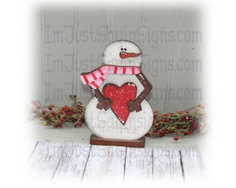 May include: Wooden snowman figurine holding a red heart with glitter. The snowman is wearing a pink and white striped scarf. The base of the snowman says "Be My Valentine".