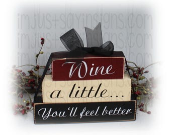 May include: A three-tiered wooden sign with the text "Wine a little... You'll feel better". The top tier is burgundy red, the middle tier is cream, and the bottom tier is black. The sign is decorated with a black bow.