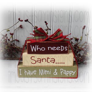 May include: A three-tiered wooden block sign with a red and green plaid bow. The top block reads "Who needs Santa...", the middle block reads "Santa...", and the bottom block reads "I have Mimi & Pappy".