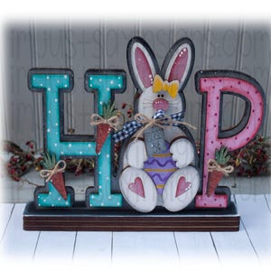 May include: A wooden sign spelling "HOP" with a bunny holding a basket of eggs. The bunny is white with pink ears and a yellow bow. The letters are painted in turquoise, pink, and white with polka dots.