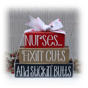 May include: Stacked wooden blocks with the text "Nurses... Fixin' cuts And Stickin' Butts" in white lettering. The top block is red, the middle block is beige, and the bottom block is black. A white ribbon with pink hearts is tied around the blocks.