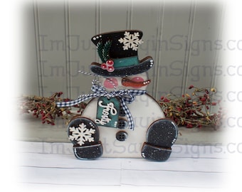 May include: A wooden snowman figurine with a black top hat, a green scarf, and a white snowflake on the hat. The snowman is wearing black mittens and has a red nose and mouth. The snowman is holding a tag that says "Frosty".