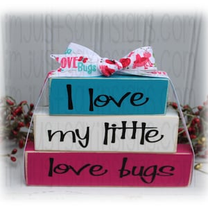 May include: A stack of three wooden blocks with the phrases "I love", "my little", and "love bugs" in black script. The top block is teal, the middle is white, and the bottom is magenta. A white ribbon with red ladybug and heart designs is tied on top.