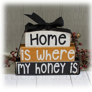 May include: A three-tiered wooden block sign with the text "Home is where my honey is" in black and white lettering on a black, orange, and white background. The blocks are tied together with a black ribbon.