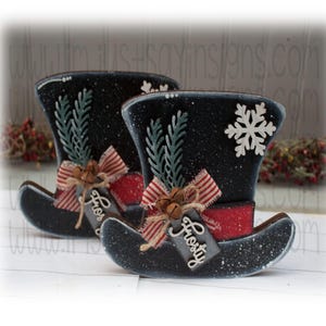 May include: Two black and white snowman hats with red and white striped ribbon, green sprigs, and bells. The hats have a tag that says "Frosty".