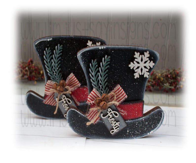May include: Two black and white snowman hats with red and white striped ribbon, green sprigs, and bells. The hats have a tag that says "Frosty".