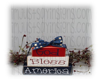 Op de afbeelding: Gestapelde houten blokken met een patriottisch rood, wit en blauw thema. De bovenste blok leest "God", de middelste blok leest "Zegen", en de onderste blok leest "Amerika". Een blauw lint met gouden sterren is om de bovenste blok gebonden.