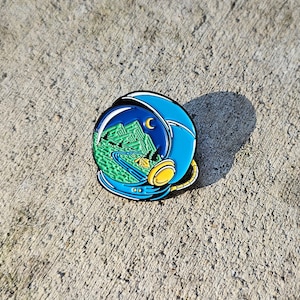 May include: A blue enamel pin shaped like an astronaut's helmet with a view of a green mountain range, a winding road, and a campsite inside. The helmet has a yellow visor and a black outline.