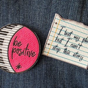 May include: Two embroidered patches on a denim background. One is a circle with a piano key design and the words "be positive." The other is a notepad-shaped patch with the text "I lost my place, but I can't stop this story."