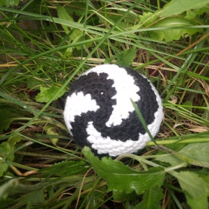 May include: A black and white crocheted ball with a swirl pattern, nestled in green grass.